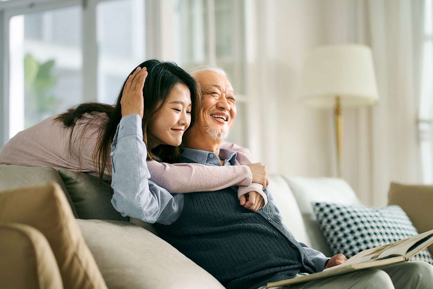 a smiling young woman hugging a smiling elderly man over the back of a couch as he reads a book
